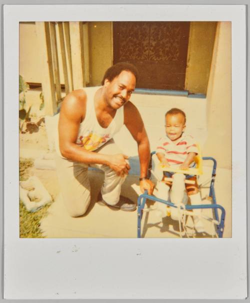 [Man in tank top crouched beside toddler on rocking horse outside]