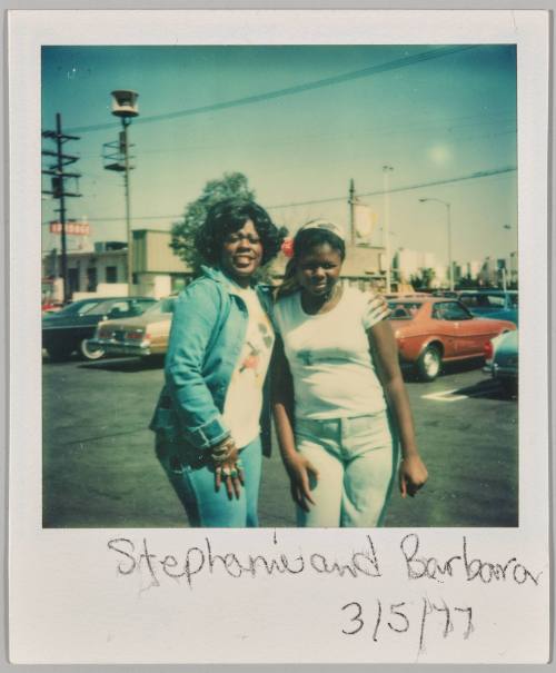 Stephanie and Barbara 3/5/77 [Two women standing in car lot, woman on left in denim jacket and has her arm around the other woman]