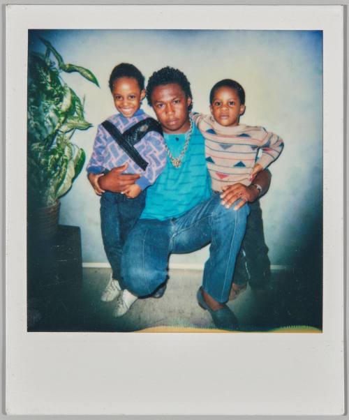 [Man with gold chain crouching with his arms around two children, house plant to left]