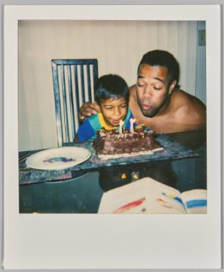 [Boy in chair and man crouching beside him, both blowing out candles on cake]
