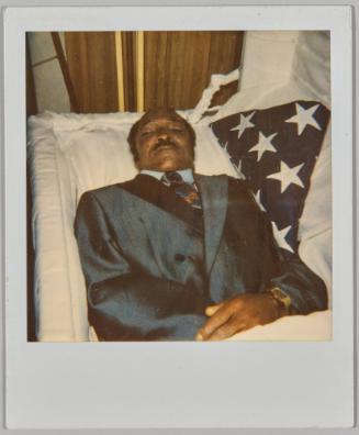 [Man in blue suit in coffin with white lining, blue fabric with white stars to right]