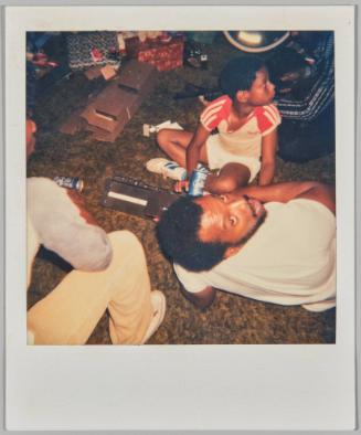 [Boy sitting cross legged on floor with man lying down with his head turned to the camera]