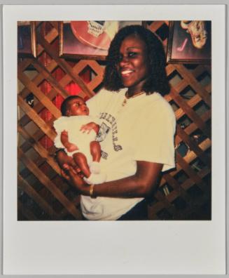 [Woman in white t-shirt holding baby, standing inside in front of wood lattice wall with framed pictures]