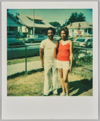 [Man in white t-shirt and woman in red tank standing together on lawn with chainlink fence]