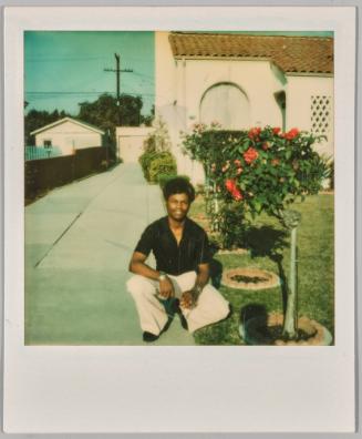 [Man in black shirt and white pants crouching in driveway beside flower bushes]