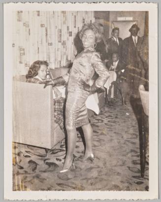 [Woman with one hand on her hip and one on the arm of a couch where another woman is sitting, "Ladies Room" sign in background]