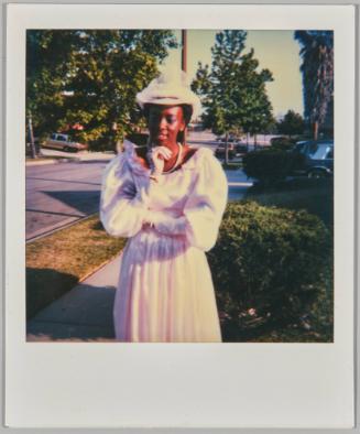 [Woman in white dress, gloves and hat standing on sidewalk, she has her hand on her chin]