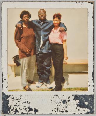 [Man with arms around two women standing in front of wooden bench and white wall]
