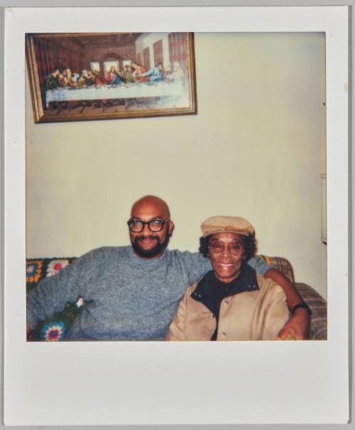 [Man wearing black rimmed glasses sitting on couch with arm around elderly woman wearing brown hat]