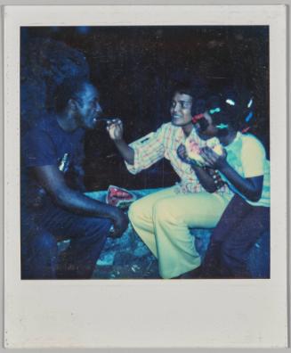 [Man and woman sitting together, woman is eating a piece of watermelon, man has one hand raised]