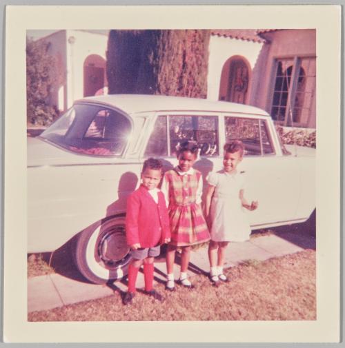 [Two young girls and a boy dressed formally standing on lawn beside driveway in front of a white car]