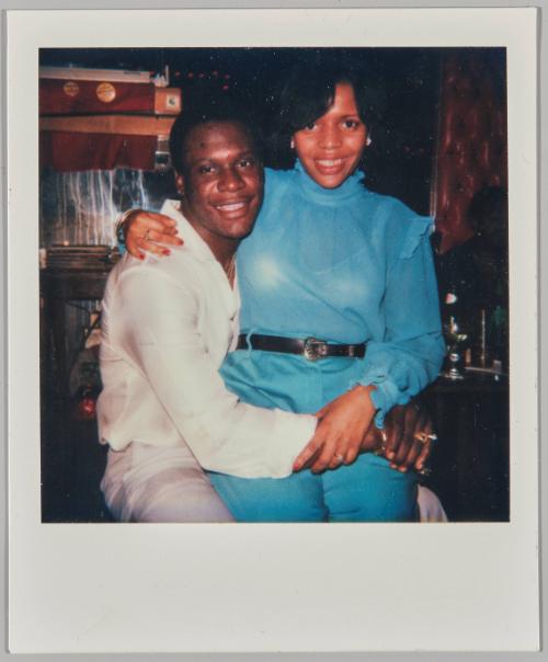 [Woman in blue blouse and blue pants sitting on man's lap, both smiling]