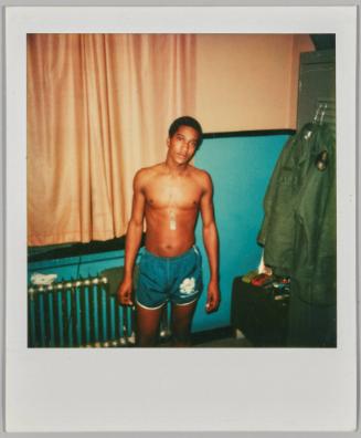 [Young shirtless man standing by window wearing turquoise shorts]