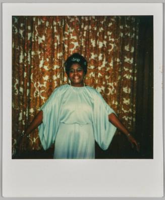 [Woman in light blue gown and hair accessories posing in front of patterned drap with her arms out on either side]