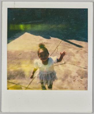 [Toddler girl in ruffled skirt standing on sidewalk with hand up]