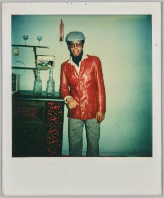 [Man in red leather jacket and sunglasses leaning against red patterned mantel]