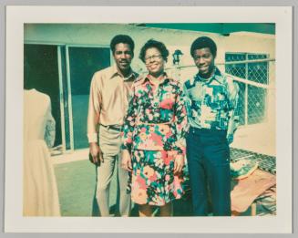 [Woman in floral dress standing inbetween two men in button up shirts, sliding door in background]

