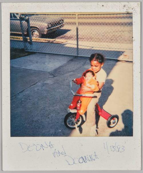 Deona and [Deanisa?] 1/13/83 [Little girl on red tricycle and holding a baby in orange onesie, they are on pavement by chainlink fence]