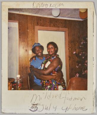 Living Room/ Midred Fatman 5 July at home [Man in blue hat and woman in patterned dress hugging in a wood panneled room with a white door]