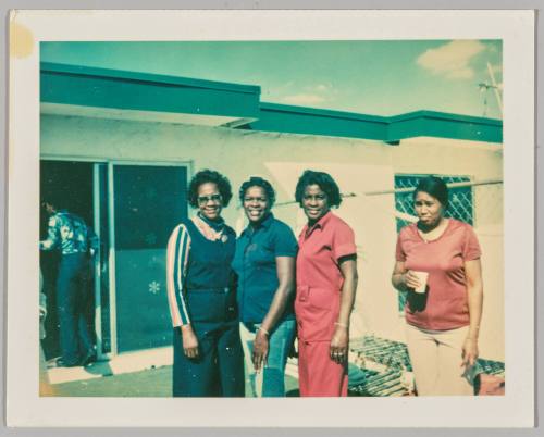 [Three women standing together outside and smiling, another woman standing off to the right and pouting with a cup in her hand, man walking through sliding doors on left ]