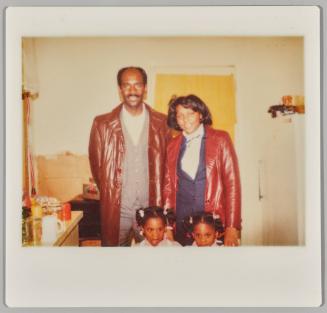 [Man and woman standing in kitchen with two girls, both wearing leather jackets]
