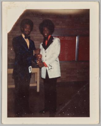 [Two men standing and holding their fists stacked on top of each other, man on right wearing white suit jacket and a sticker labeled “12” on his hip]
