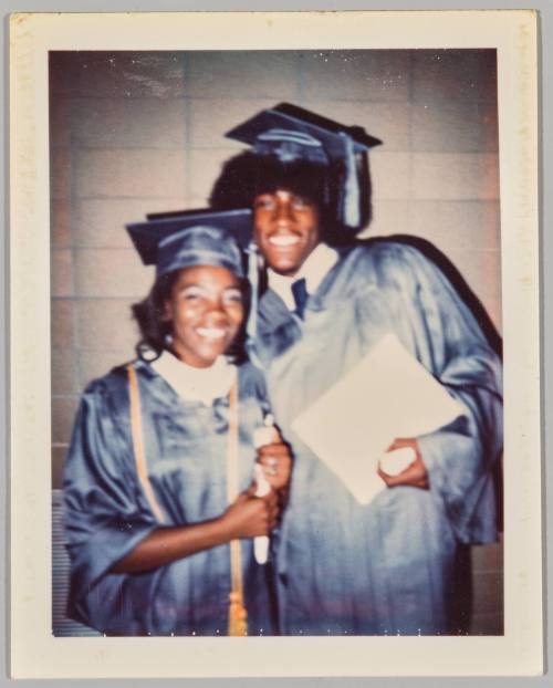 [Adolescent man and woman in graduation robes and hats, man on right holding certificate]
