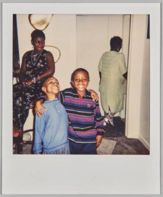 [Two boys smiling with arms around each others shoulders, two women in background, one of the women is walking into another room]
