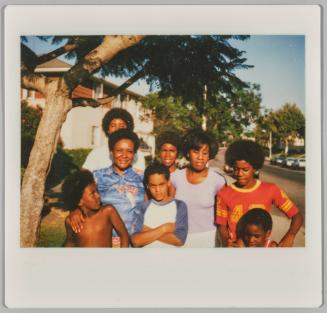 [Six boys, varying ages standing in a group with two women on street beneath a tree branch]
