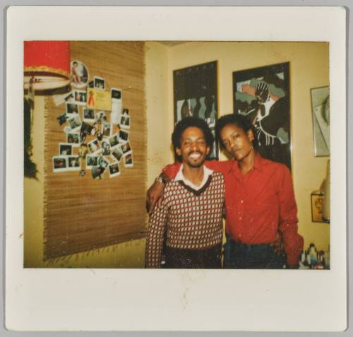 [Two men standing together, man on right has arm around man on left, next to wall with Polaroids displayed]
