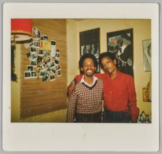[Two men standing together, man on right has arm around man on left, next to wall with Polaroids displayed]
