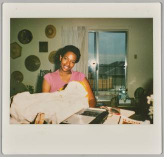 [Woman smiling with what appears to be a swaddled baby, hats are hung on wall in background on the left]
