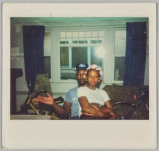 [Man sitting with woman wearing curlers in her hair on his lap with large window in background]
