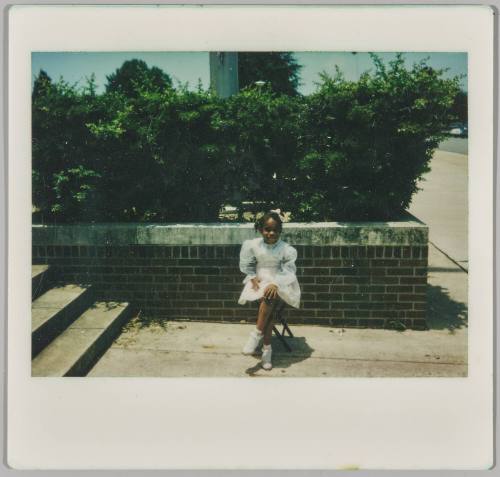 [Little girl in white dress sitting on stool outside in concrete area next to stairwell]
