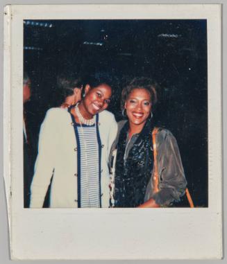 [Two women standing side by side smiling, woman on left wearing baby blue cardigan and striped dress]
