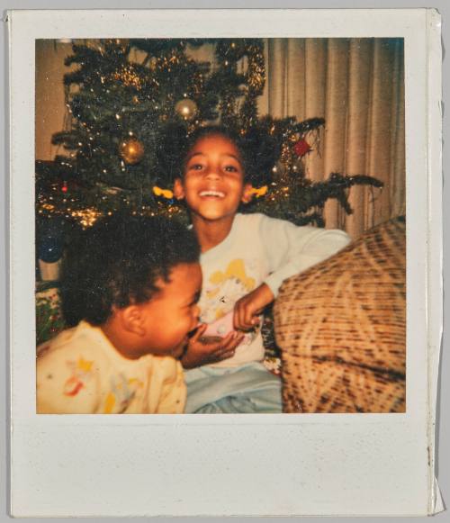 [Little girl smiling with Christmas tree behind her, toddler in foreground]
