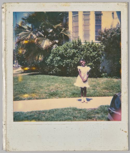 [Little girl posing in frilly white dress on sidewalk next to home with tropical plants in background]
