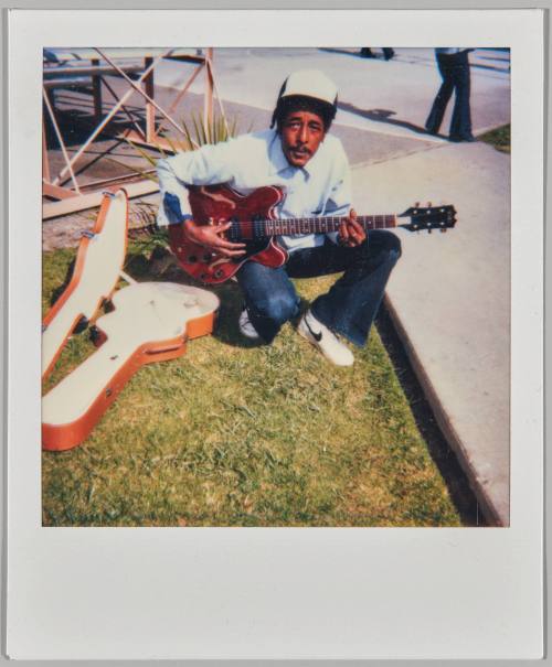 [Man sitting on grass with guitar in hands and guitar case on the left side]
