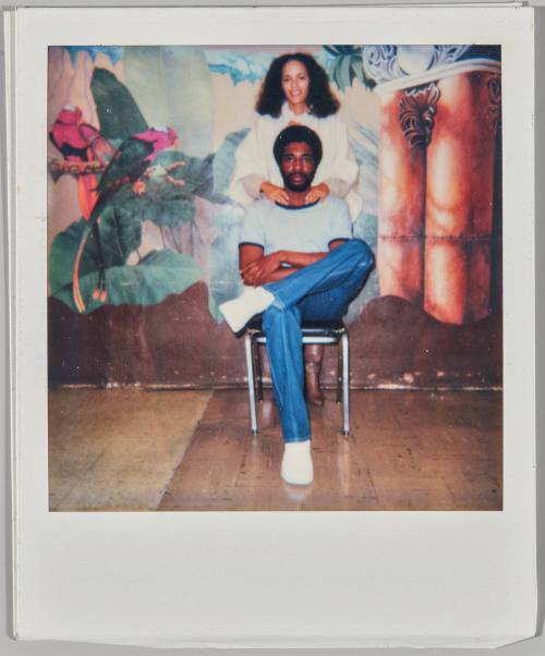 [Woman standing behind seated man with her hands on his shoulders in front of mural on back wall]
