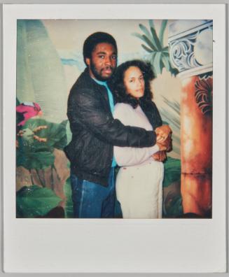 [Man embracing woman from behind, holding hands, standing in front of painted mural on wall with red bird on left side]
