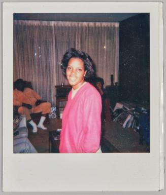 [Woman in pink sweater smiling, two people sitting in background in living room, man on left in orange outfit]
