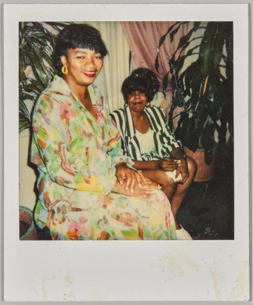 [Woman in colourful dress sitting with hands in lap with other woman in striped blazer sitting behind her]
