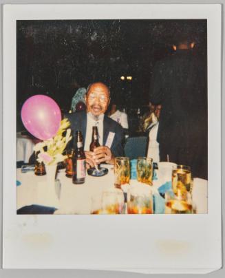 [Elderly man wearing glasses sitting at table with pink balloon and beer bottles]
