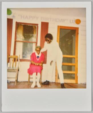 [Woman sitting and smiling in red outfit on porch with woman dressed in all white, banner above doorway stating ‘Happy Birthday Lu’]
