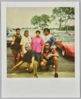 [Four women standing side by side with two men crouching in front in parking lot]
