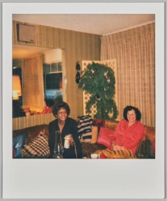 [Two women sitting on couch drinking cans of beer with plant and window to other room behind them]
