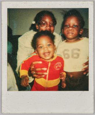 [Girl holding two toddlers in hands, toddler in front is smiling, toddler to the right wearing a shirt with ’66’ on the front]
