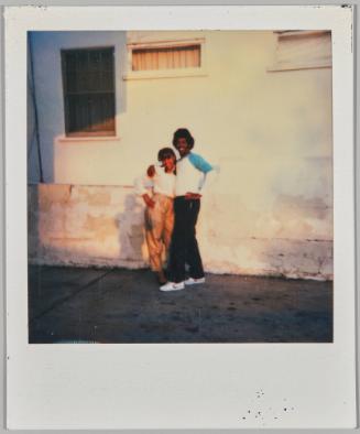 [Man and woman with arms around each other smiling with hands on hips against white wall]
