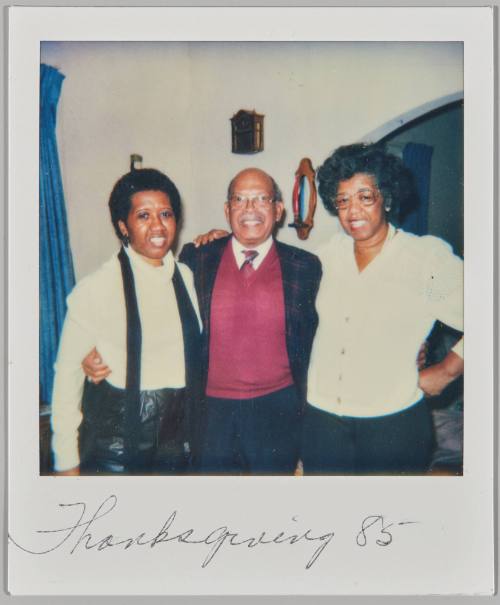 Thanksgiving '85 [Man in burgundy sweater vest posing with women on either side of him in home]
