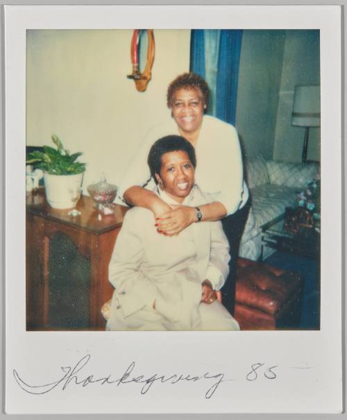 Thanksgiving '85 [Woman sitting on chair and smiling while other woman hugs her around the shoulders from behind]

